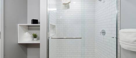 Une salle de bain moderne avec une douche à parois de verre, des murs recouverts de carreaux blancs et une étagère où sont posées des serviettes et une plante.