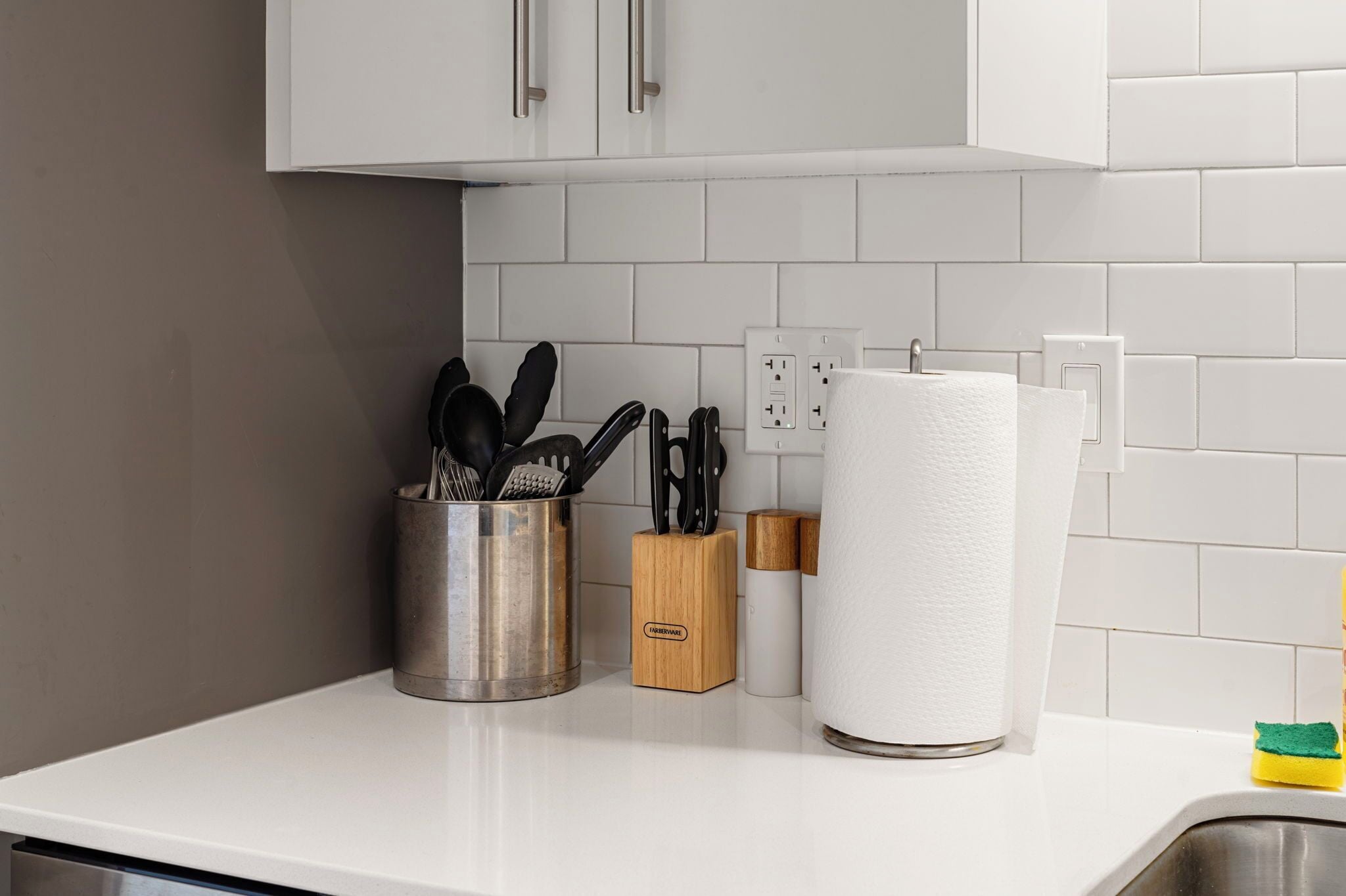 Una cocina con armarios blancos, un recipiente de acero inoxidable para utensilios, un bloque de madera para cuchillos, un dispensador de toallas de papel y una esponja amarilla.
