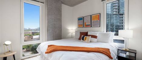 A modern bedroom with a large bed, two windows, and artwork on the wall.
