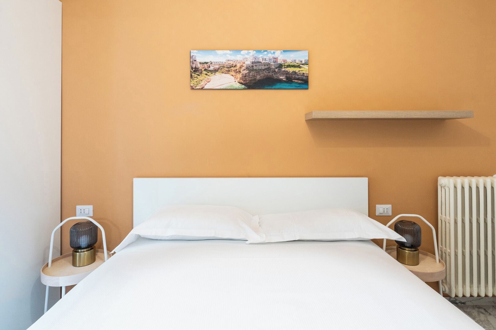 A hotel room with a bed, a painting of a coastal town, a bedside table with a lamp, and a radiator.