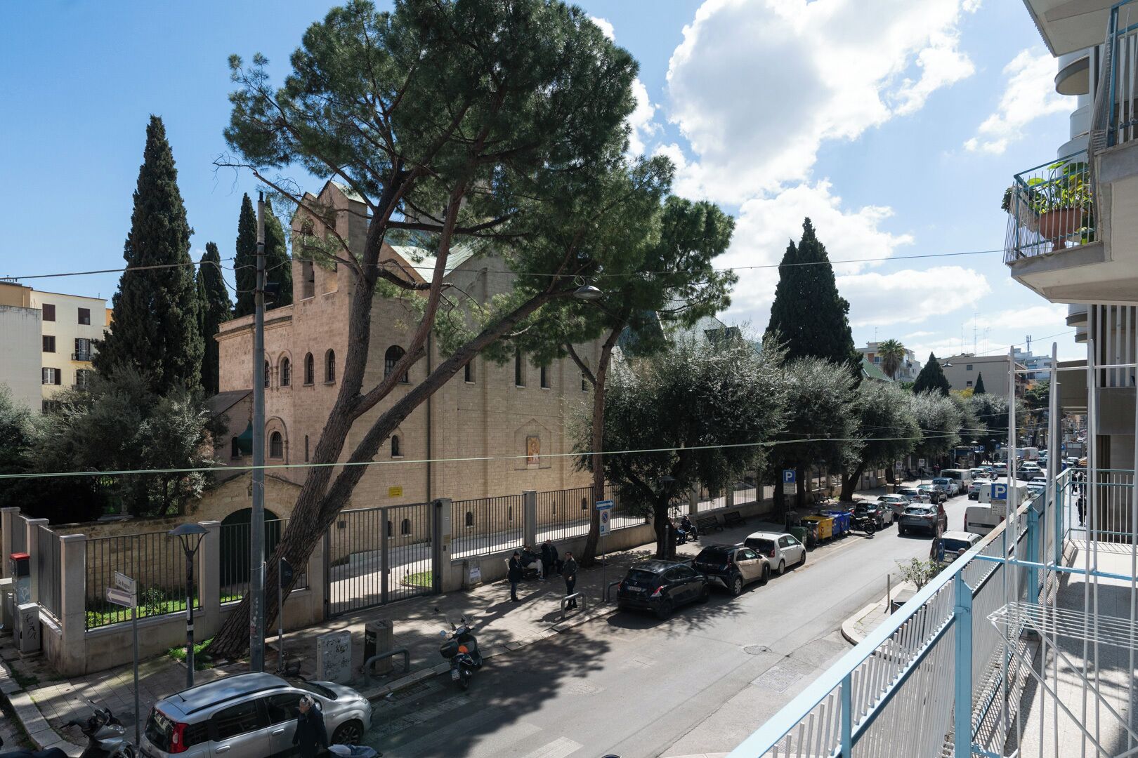 A street view with parked cars, a building with a tower, and trees.