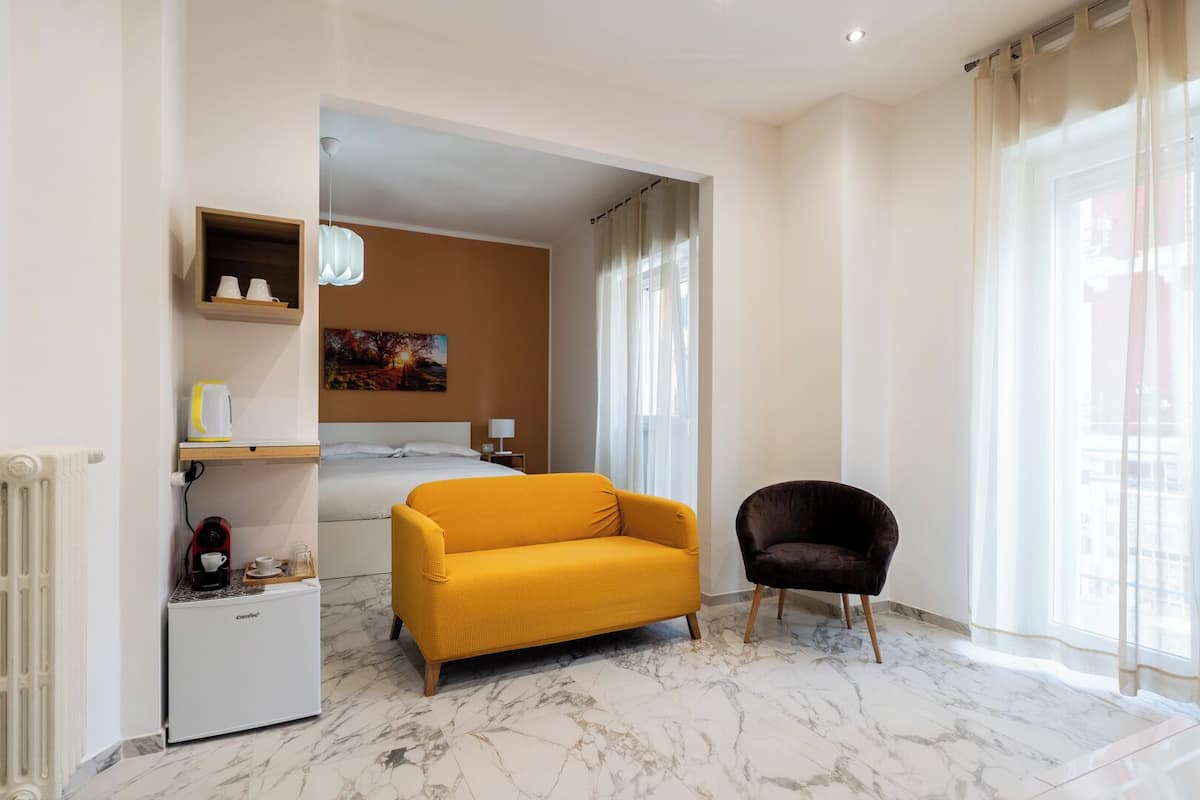 A modern hotel room with a marble floor, a yellow sofa, a dark armchair, a small kitchenette, and a bed with a painting on the wall.