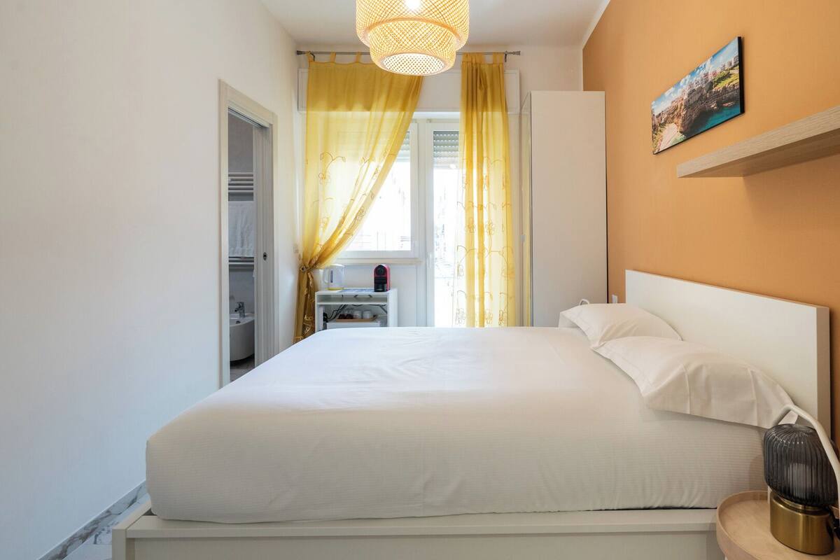 A hotel room with a bed, a bedside table with a lamp, a small table with a kettle and a red device, and a painting on the wall.