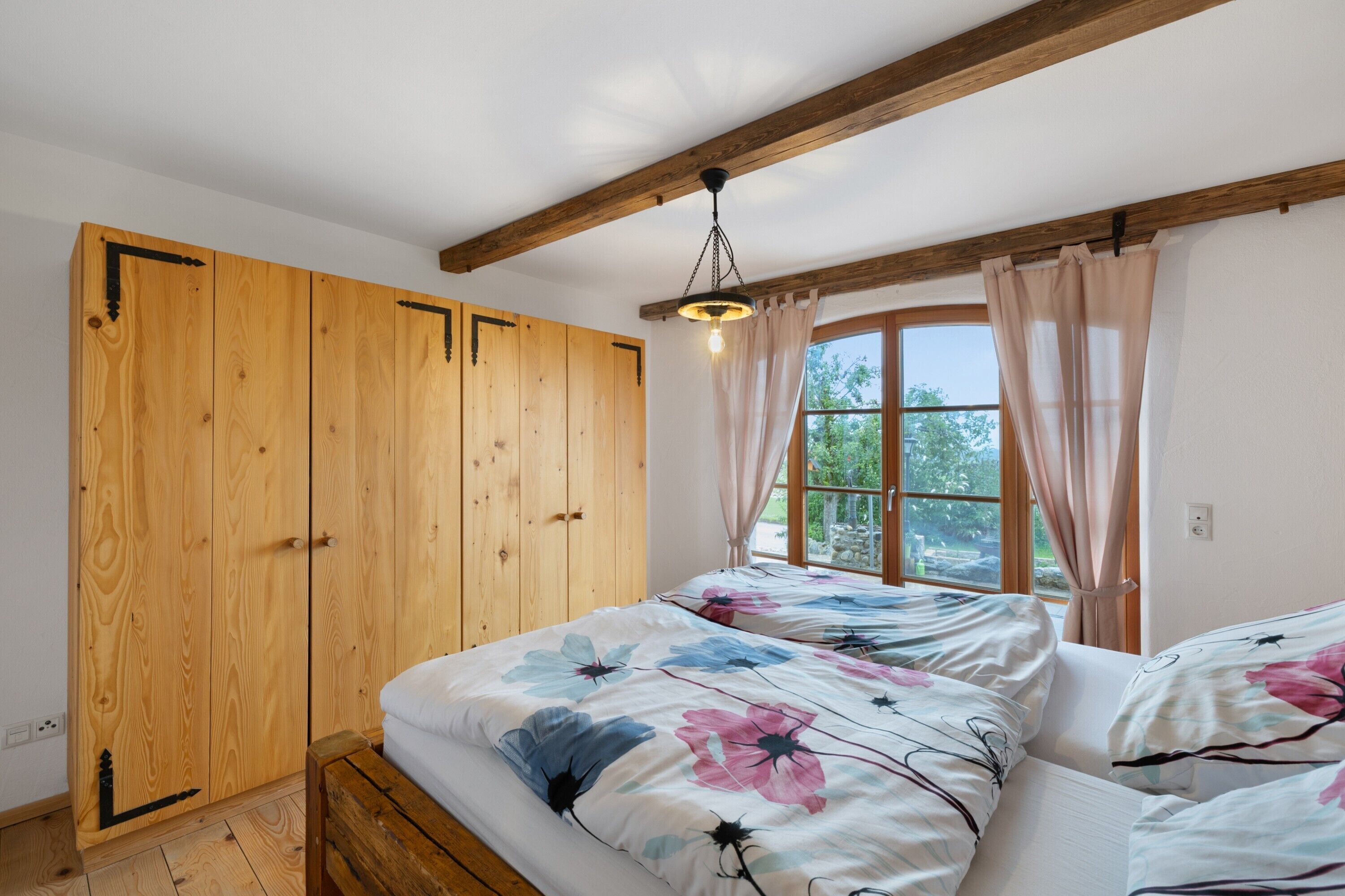 A bedroom with wooden wardrobe, floral bedding, and a window with curtains.