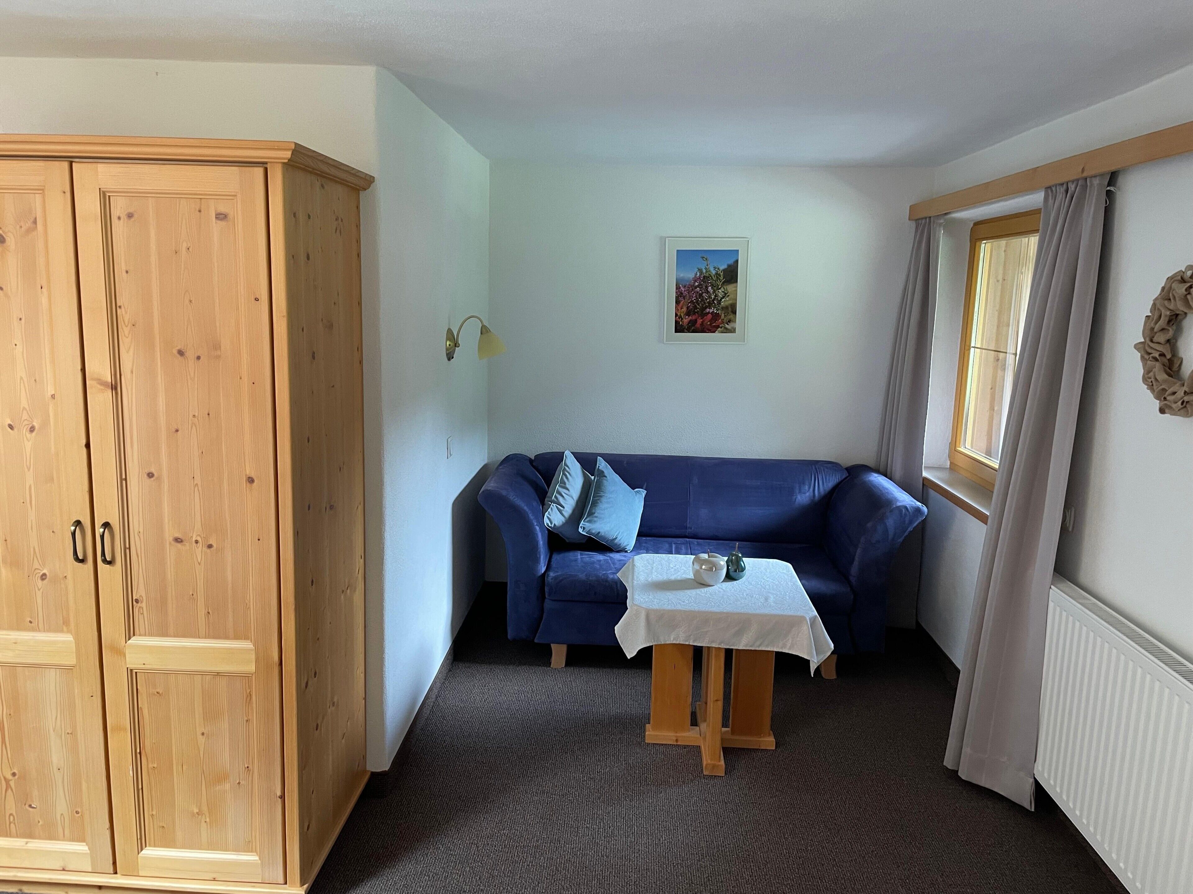 Een kamer met een blauwe bank, een houten garderobe, een klein tafeltje met een wit tafelkleed en een raam met gordijnen.