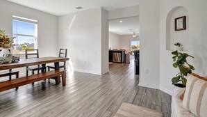 Amplia sala de estar con una mesa de madera y bancos, una cocina al fondo y un gran ventanal que proporciona luz natural.