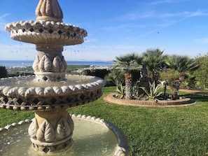 A stone fountain with water spouting from its top, surrounded by a circular brick border and a grassy area with palm trees. - Apartment 'Villa Roca' with Sea View, Wi-Fi and Air Conditioning (Málaga)