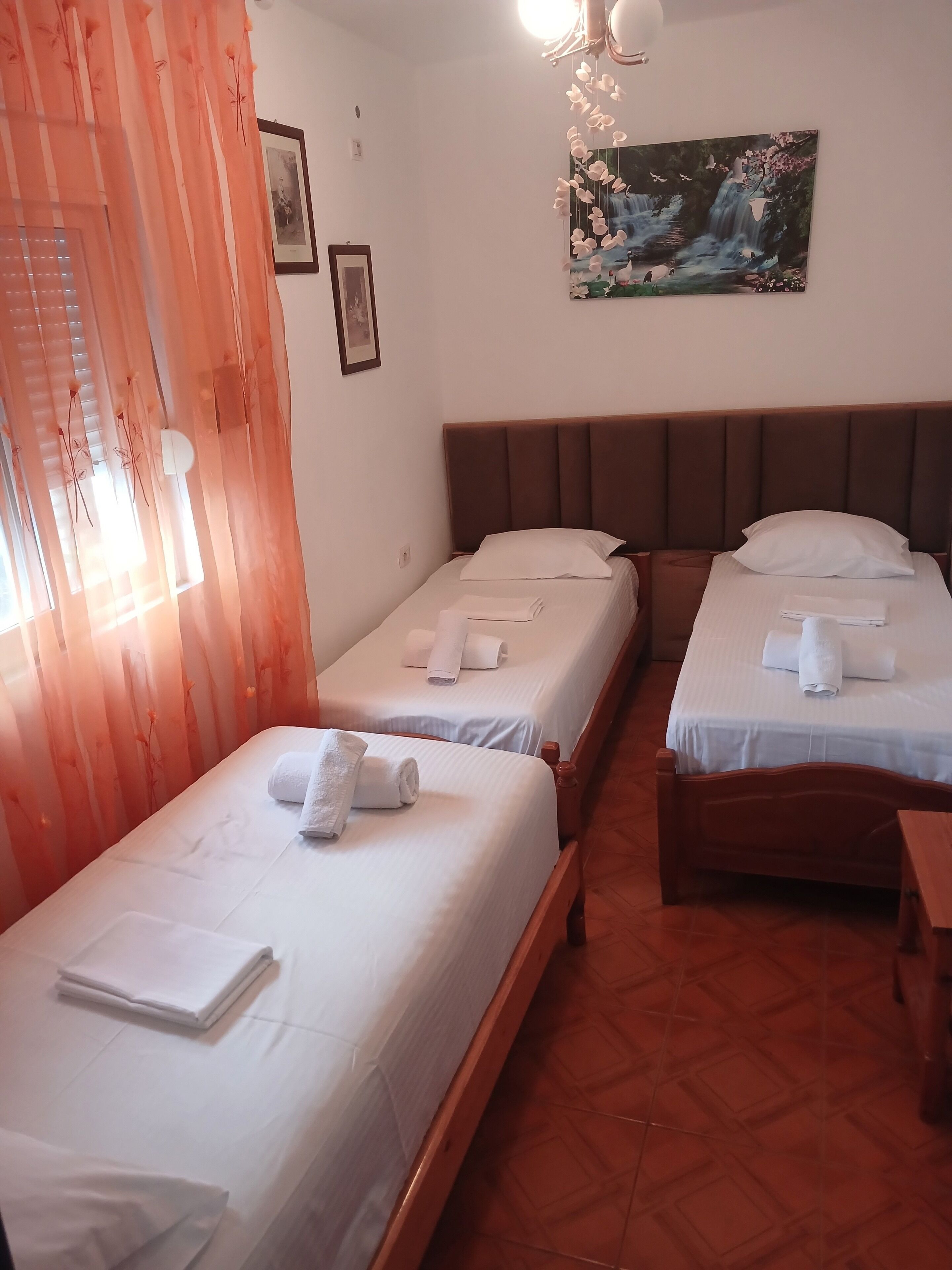 A hotel room with two single beds, each with a pillow and a rolled towel. There is a painting on the wall, a window with curtains, and a wooden floor.