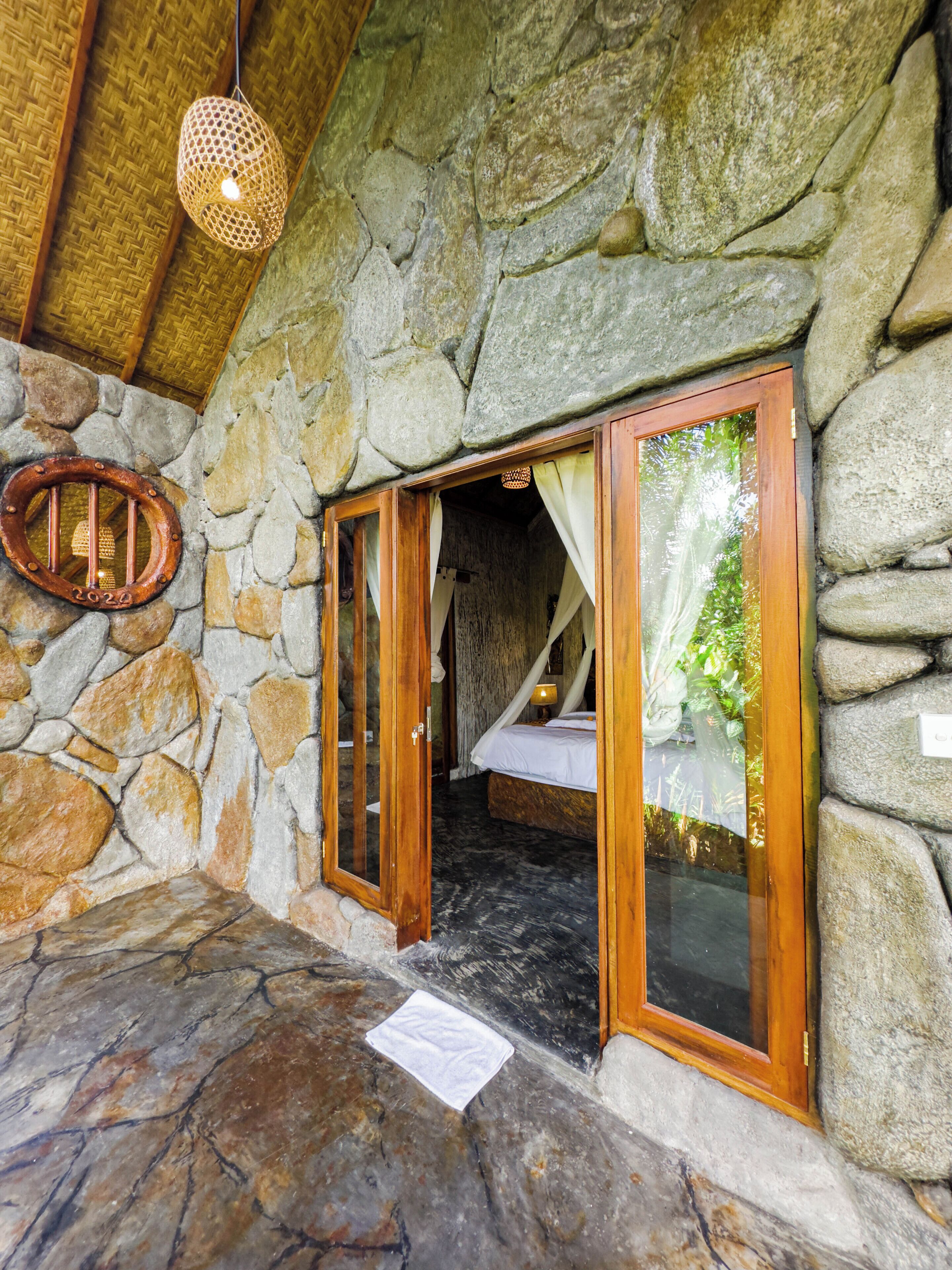A stone-walled room with a bed, a hanging light fixture, and a wooden door.