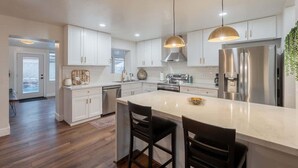 A modern kitchen with white cabinets, stainless steel appliances, and a central island with bar stools. - Spacious 5BR Retreat + Game Room (Phoenix)