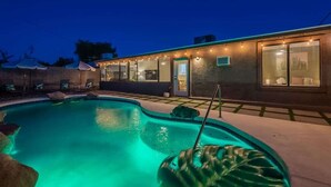 A swimming pool with a stone accent wall, a covered patio area with chairs, and a building with a covered entryway. - Spacious 5BR Retreat + Game Room (Phoenix)