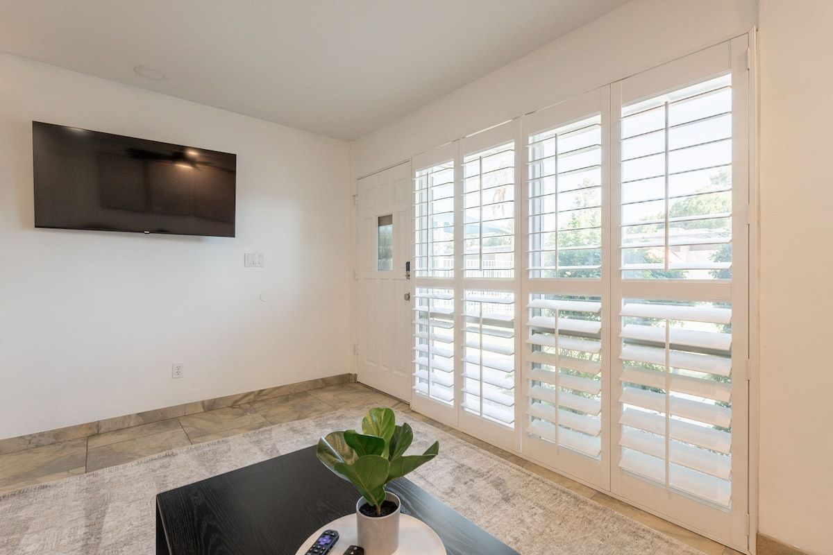 A room with a flat-screen TV mounted on the wall, a potted plant on a table, and large windows with blinds.