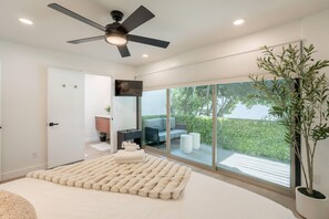 A bedroom with a ceiling fan, a large window, a bed with a fluffy rug, a TV, and a potted plant.