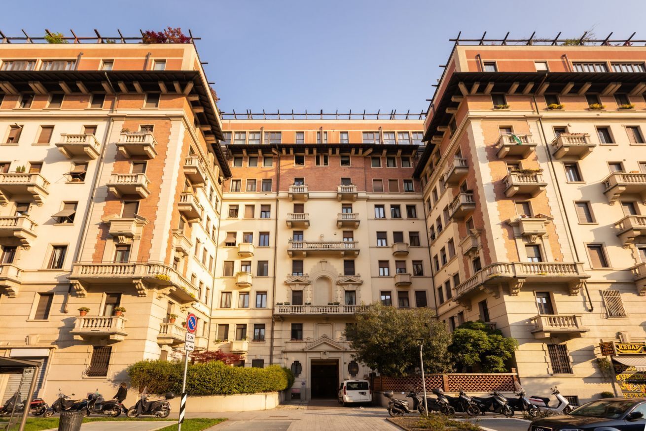 A multi-story building with a central entrance, balconies, and a parking area with scooters.