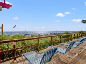 A wooden deck with blue chairs, a railing, and a view of the beach with umbrellas and the ocean.