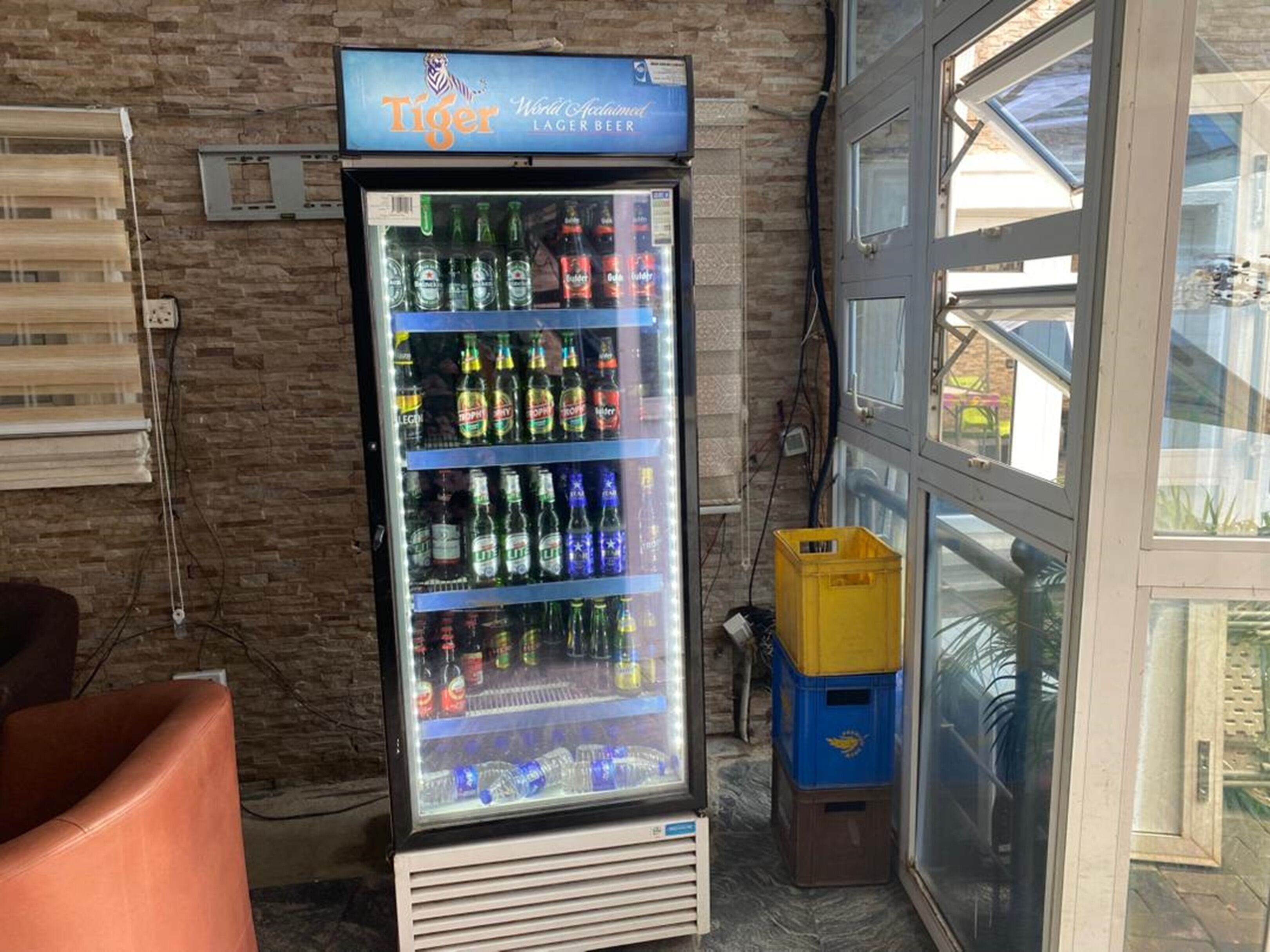 Un refrigerador lleno de diversas bebidas, incluyendo cerveza y agua, en una habitación con pared de ladrillo y ventana.