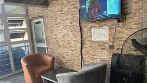 Egy modern, kényelmes pihenősarok, melyben egy bőrkarosszék, egy szürke fotel, egy kis asztal, egy téglás falra szerelt televízió és egy állóventilátor található.