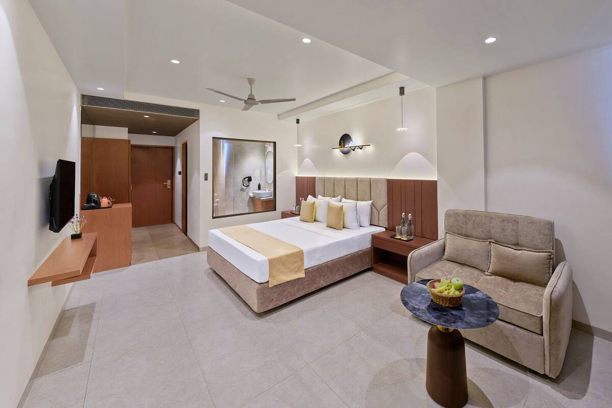 A modern hotel room with a large bed, a sofa, a small round table, a TV, and a mirror.