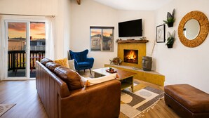 A living room with a sofa, a blue armchair, a coffee table, a fireplace, a television, and a balcony with a view.
