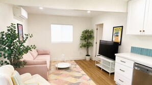 A living room with a sofa, a coffee table, a TV, a kitchen area with a dishwasher, and a window. - Walk To Beach, Surfing, Mins 2 Restaurants & Shops (Oxnard)
