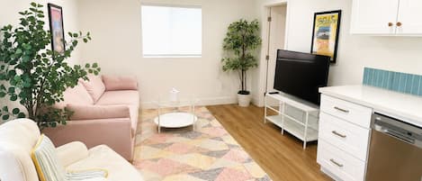 A living room with a sofa, a coffee table, a TV, a kitchen area with a dishwasher, and a window.