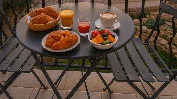 A table set with croissants, fruit, juice, and coffee on a balcony overlooking a cityscape at sunset.