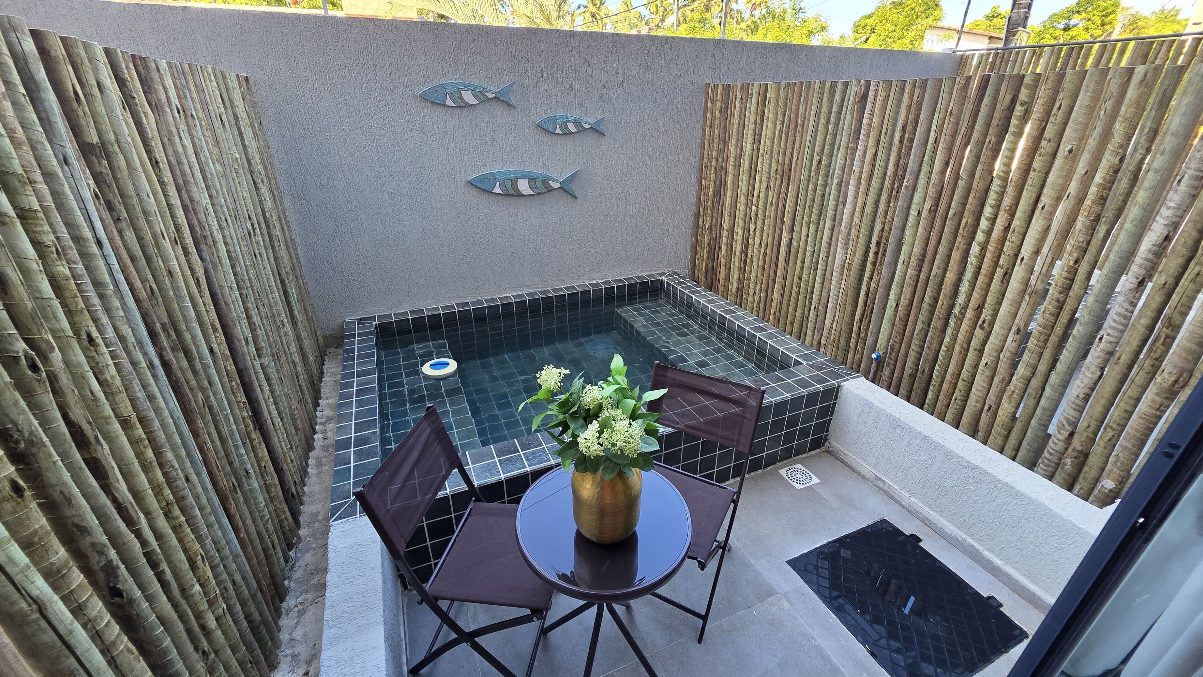 A small outdoor hot tub area with a tiled floor, surrounded by bamboo fencing. There are three fish-shaped decorations on the wall above the tub.