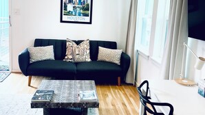 Un salon avec un canapé noir, une table basse, un téléviseur et une fenêtre avec des rideaux.