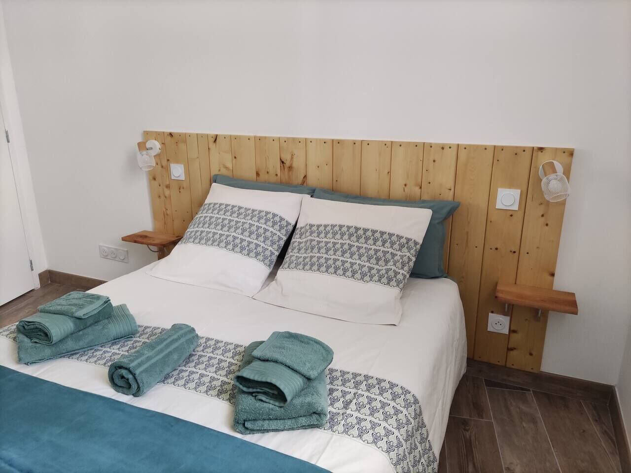 Een net opgemaakt bed met patroon kussens en gevouwen handdoeken, een houten hoofdbord en nachtkastjes met lampen.