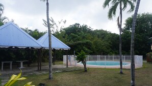 Ett poolområde med en pool som är omgiven av ett staket, palmer och en möblerad, överbyggd sittplats.