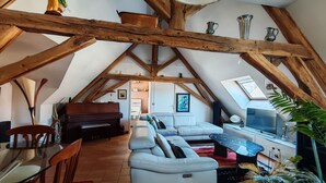 Habitación en ático con vigas de madera a la vista, un piano, un sofá blanco, un televisor y una mesa de comedor con sillas.