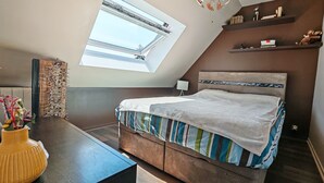 Un dormitorio con techo inclinado, tragaluz, cama con ropa de cama a rayas, mesita de noche, lámpara y estante con libros y objetos decorativos.