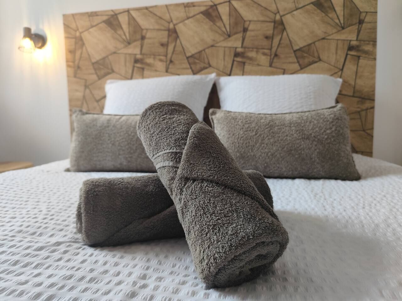 Un lit bien fait, avec des serviettes grises et des oreillers blancs, une tête de lit en bois et un luminaire fixé au mur.