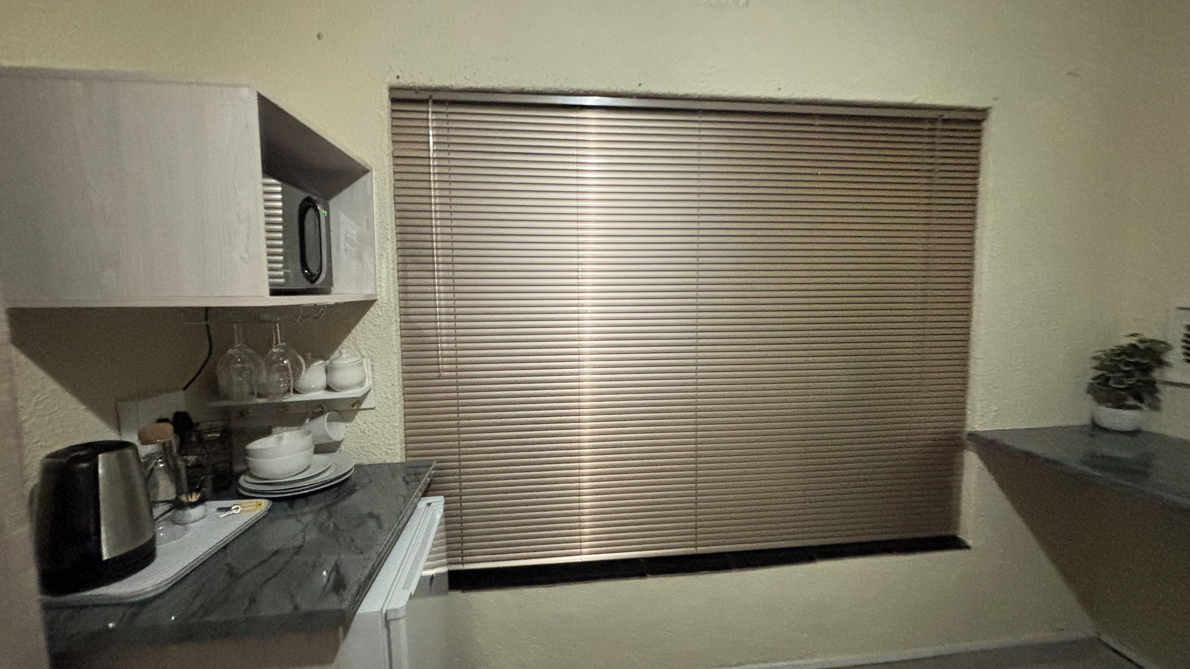 Eine kleine Küche mit Mikrowelle, Regalen mit Geschirr und einem Fenster mit Jalousien.