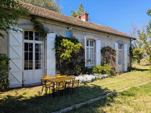 A rustic building with a tiled roof, white shutters, and a garden with a wooden table and chairs.