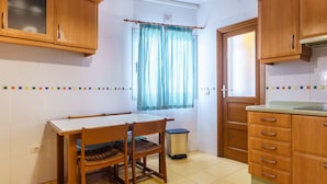 A kitchen with wooden cabinets, a tiled floor, a window with a checkered curtain, and a trash bin.