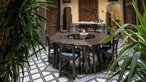 Een traditioneel Marokkaans interieur met een betegelde vloer, een centraal eetgedeelte met een ronde tafel en stoelen, en versierde houten deuren en balkons.