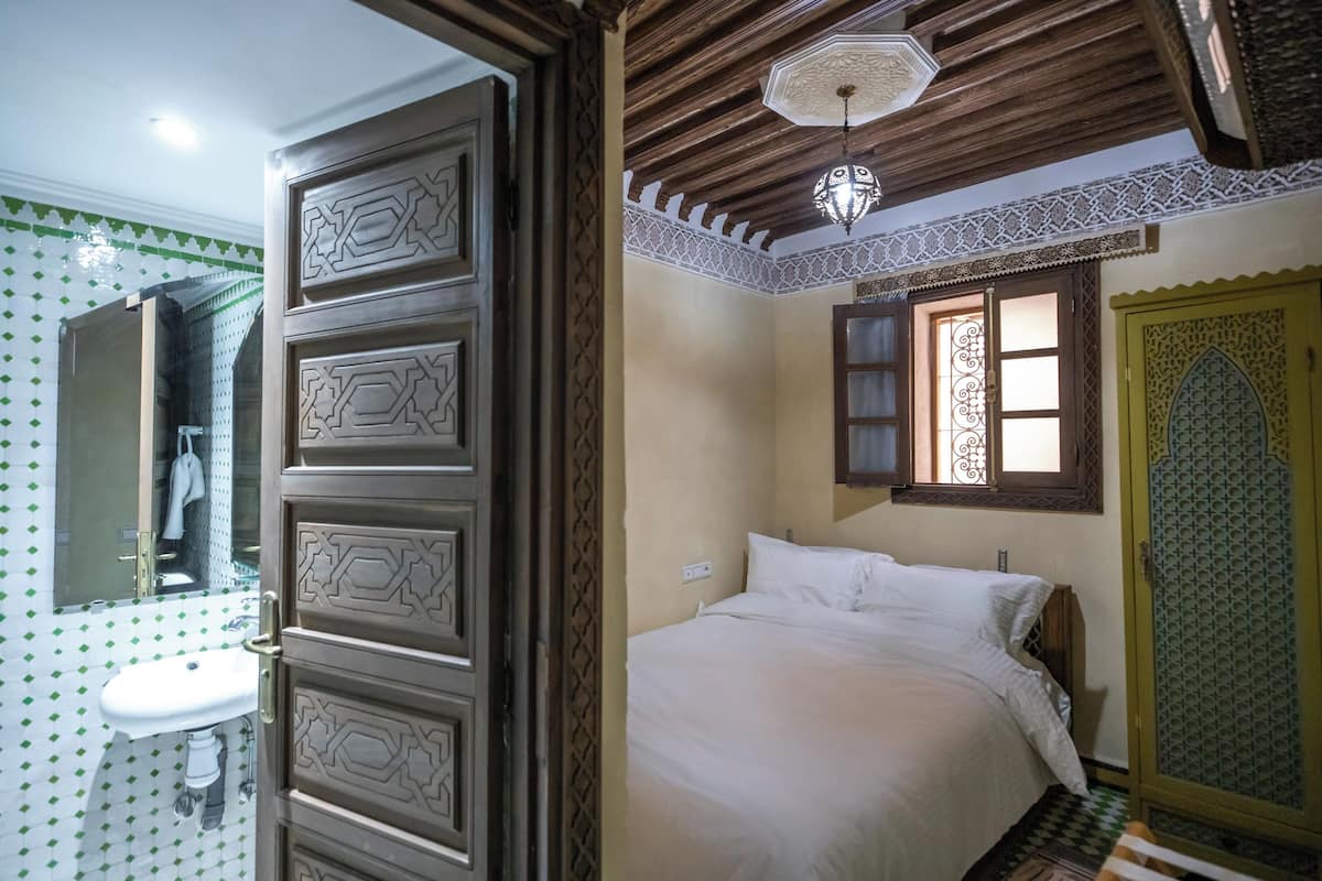 A bedroom with a bed, a window with decorative iron bars, a door with intricate patterns, and a ceiling fan.