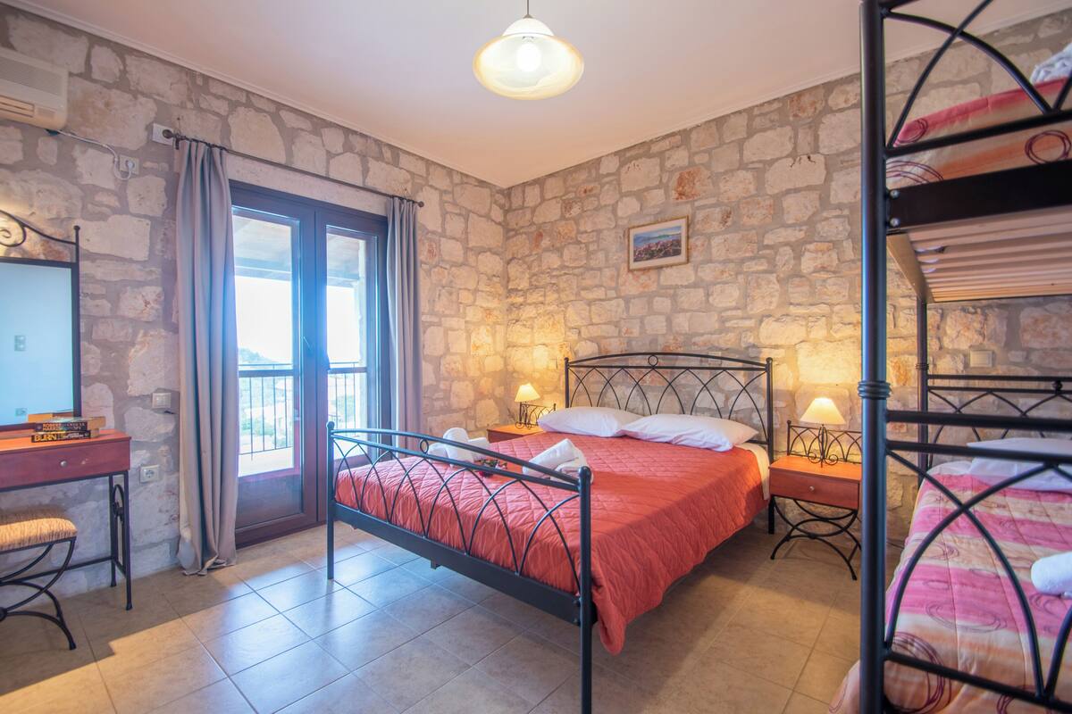 A bedroom with a stone wall, a bed with a red bedspread, a desk with a chair, and a window with curtains.