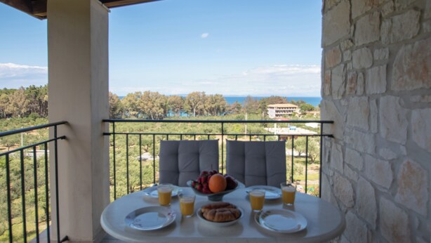 Un balcon avec une table dressée pour le petit-déjeuner, offrant une vue panoramique sur un paysage verdoyant, avec des arbres et une vue lointaine sur la mer.