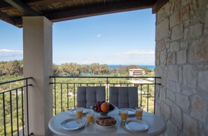 Un balcón con una mesa dispuesta para el desayuno, con vistas a un paisaje pintoresco con árboles y el mar a lo lejos.