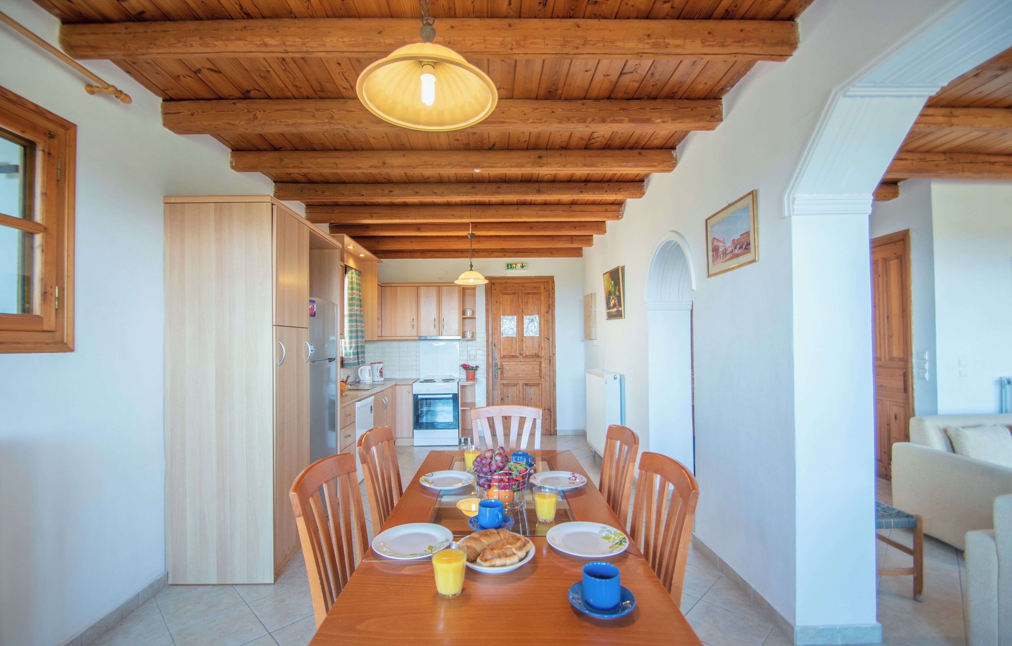 Un comedor con una mesa de madera dispuesta para cuatro personas, rodeada de sillas. Se ve una cocina al fondo con armarios de madera y un lámpara colgante.