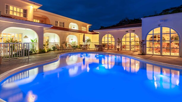 A pool area with a curved pool, outdoor seating, and a building with arched windows and warm lighting. - Santé Vivre L'Experience Livá OFICIAL (Búzios)