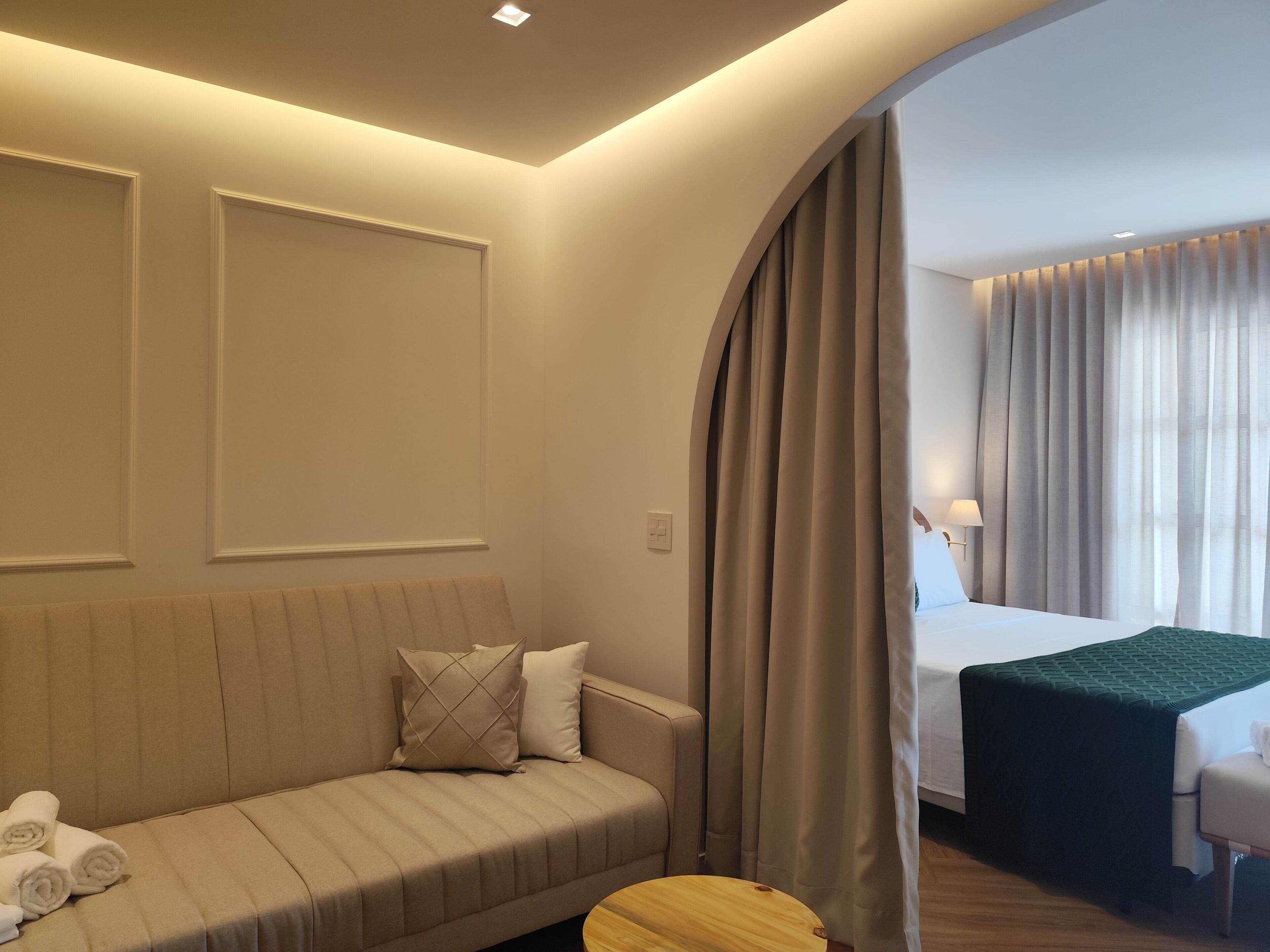 A hotel room with a sofa, a small wooden table, a bed with a green bedspread, and a window with curtains.