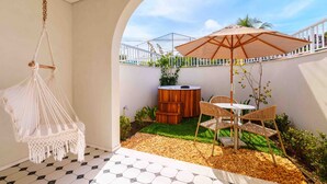 A cozy patio area with a hanging macrame chair, a wooden hot tub, and a table with chairs under a large umbrella. - Santé Vivre L'Experience Livá OFICIAL (Búzios)