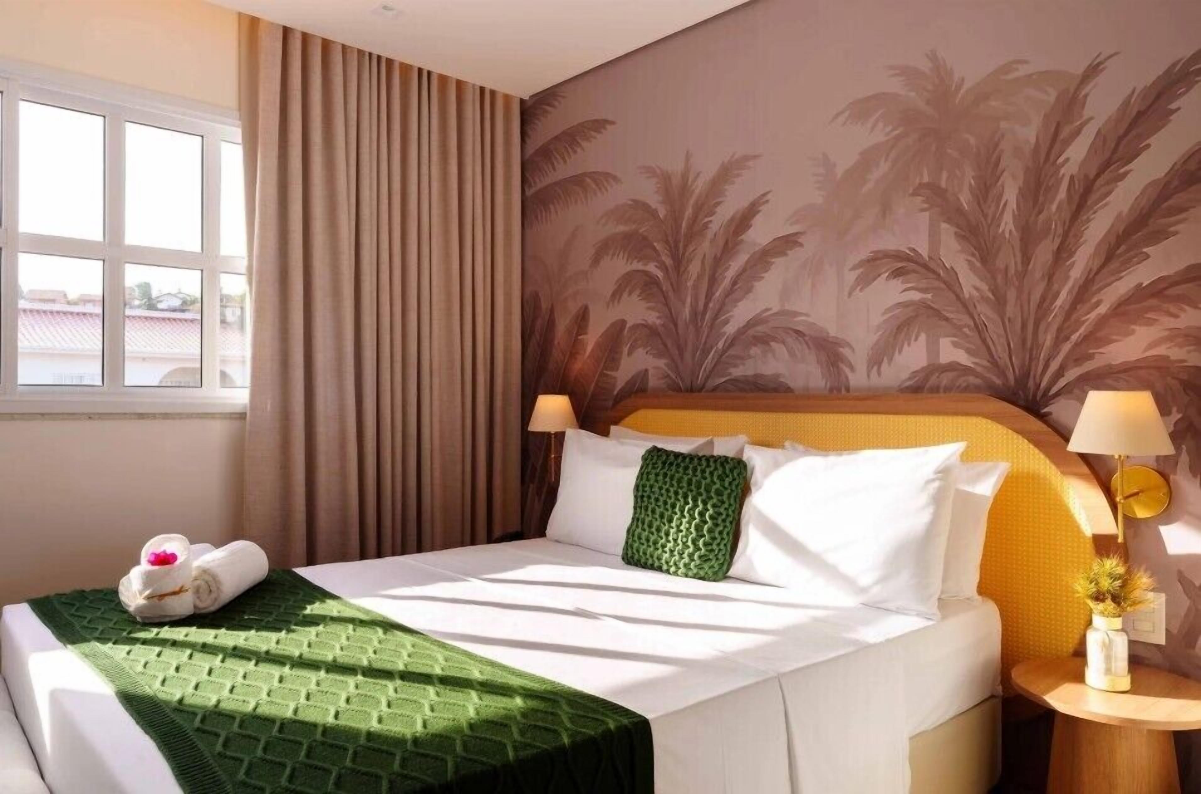 A hotel room with a bed, a window, curtains, a wall mural, and a bedside table with a lamp.