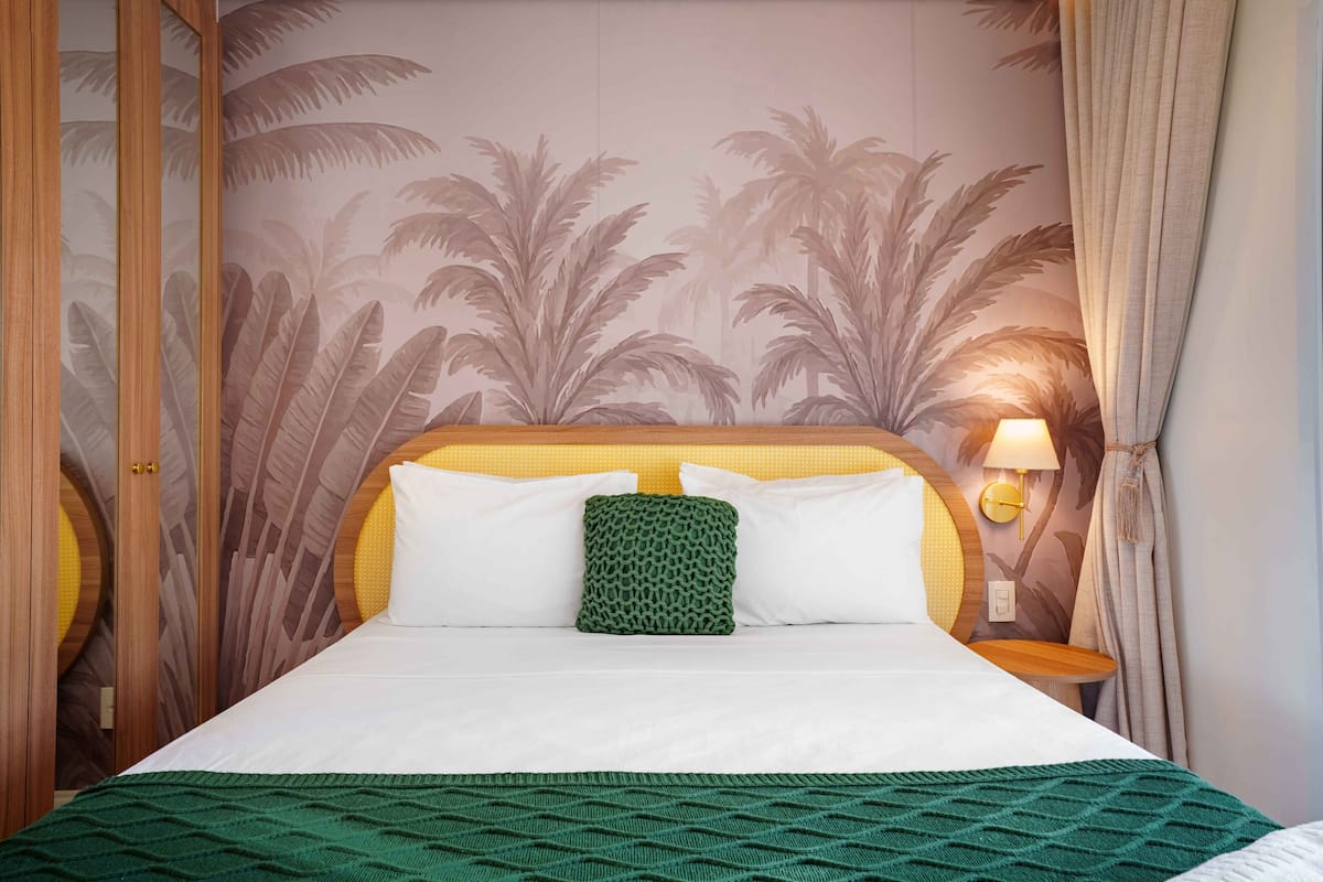 Ein Hotelzimmer mit Bett, einer grün-weißen Bettdecke, einem grünen Kissen, einer Wand mit Palmen-Tapete, einem Holz-Kopfteil und einem Nachttisch mit Lampe.