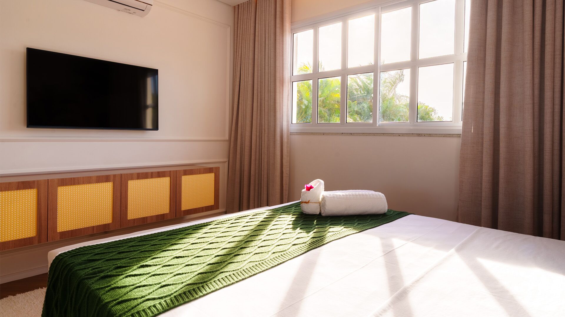 A hotel room with a bed, a flat-screen TV mounted on the wall, a window with curtains, and a wooden cabinet with a yellow panel.