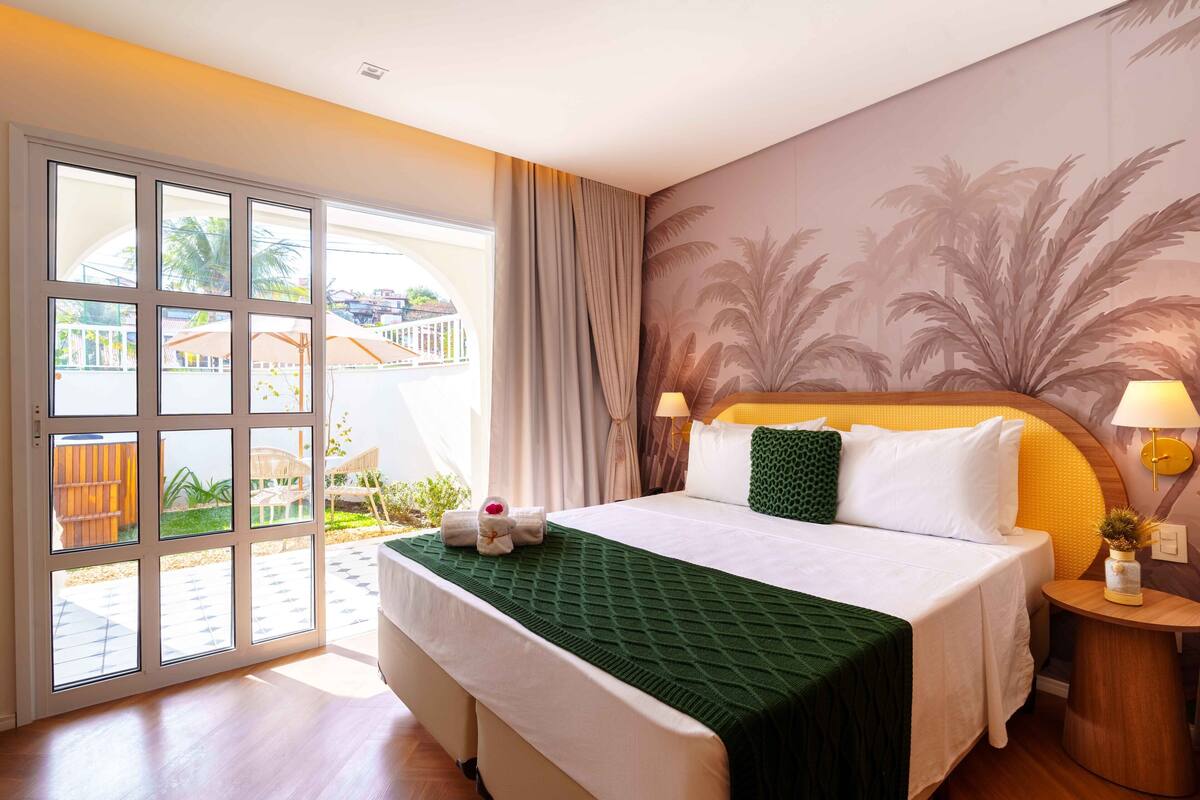 Ein Hotelzimmer mit einer großen Schiebeglasdoor, die auf einen Balkon führt, einem Bett mit einer grünen Decke, einem Kopfteil mit Palmblatt-Tapete und einem Nachttisch mit einer Lampe.