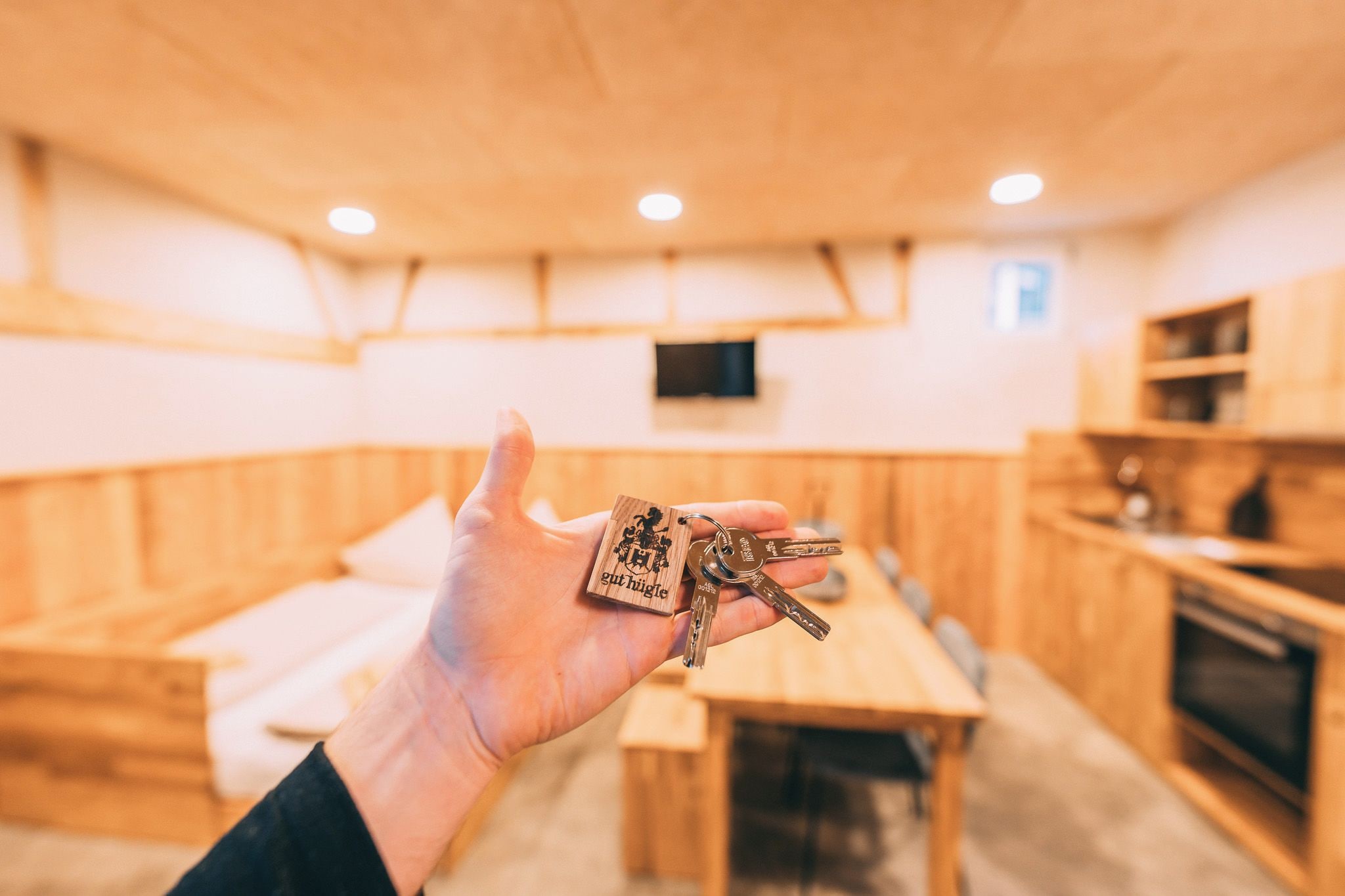 Una mano sostiene un juego de llaves con un llavero de madera que dice 'gut hicle' en una habitación con paredes de madera y una zona de cocina.